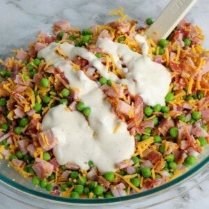 ranch dressing poured over peas, ham, bacon bits, cheddar cheese and raw onion mixed together in a bowl