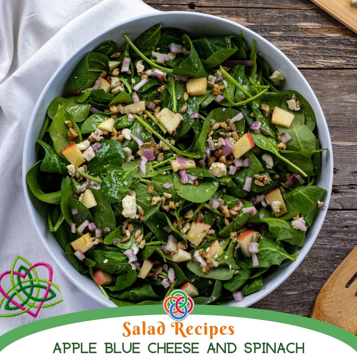 Bowl of fresh spinach salad with diced apples, blue cheese crumbles, red onion, and seeds, lightly dressed and served in a white bowl.