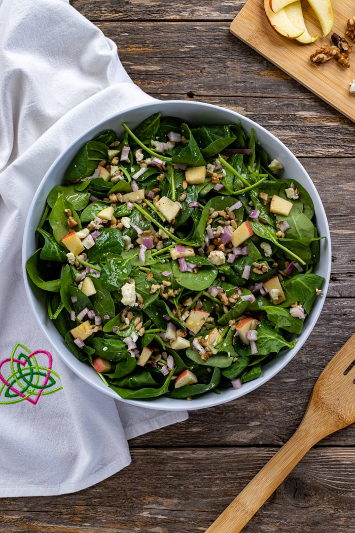 Sliced apples with spinach, blue cheese, shallots and sun flower seeds | Irish American Mom Oveerhead shot of spinach and apple salad in a bowl with a dish cloth and wooden fork