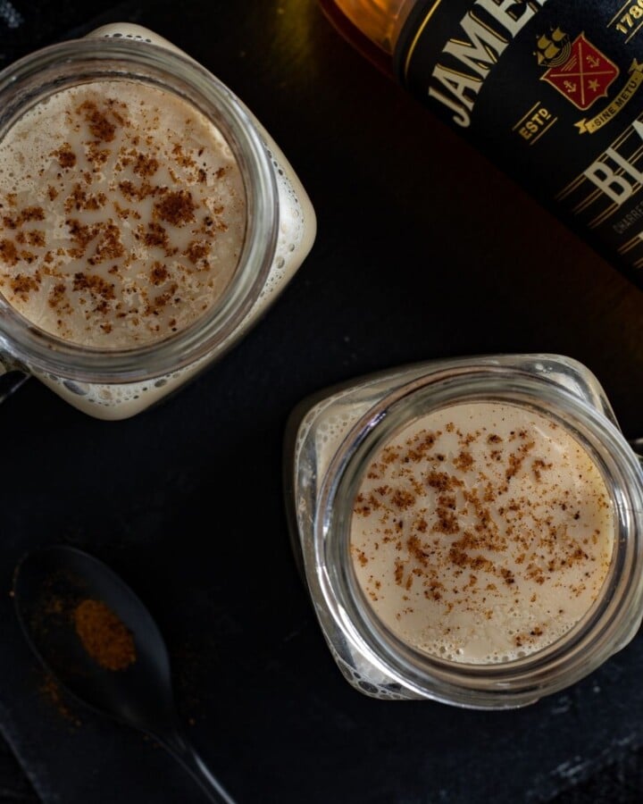 Overhead shot of milk punch in glasses with nutmeg garnish beside a bottle of whiskey