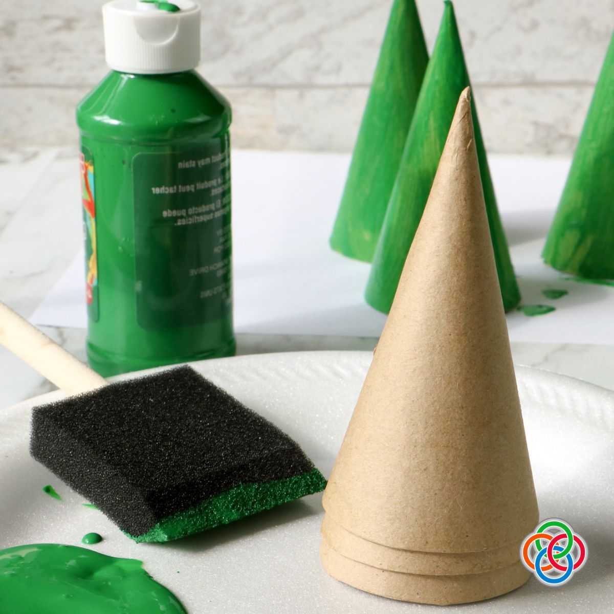 Painting cardboard cones green to make a Christmas tree craft | Irish American Mom Cardboard craft cones with a bottle of green paint and foam brush