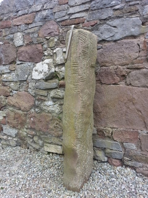 ø | Irish American Mom Standing stone with lines and indentations on its edges in front of a stone wall.