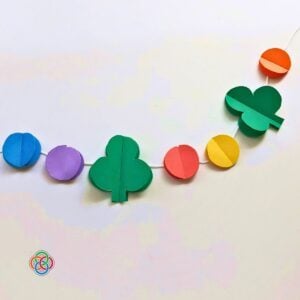 Completed Saint Patrick's Day paper garland with alternating 3D green shamrocks and colorful rainbow paper balls attached to white string, displayed against a white background with Irish American Mom logo in the corner.