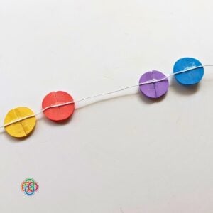 Yellow, red, purple, and blue 3D paper balls glued onto white string to form a Saint Patrick's Day garland, shown against a white background with Irish American Mom logo in the corner.