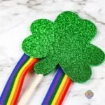 Attaching a second shamrock to a stick with ribbon strips for a shamrock wand craft | Irish American Mom Close-up of a glittery green foam shamrock being attached on top of ribbon streamers and a wooden dowel to finish a Saint Patrick's Day shamrock wand craft.
