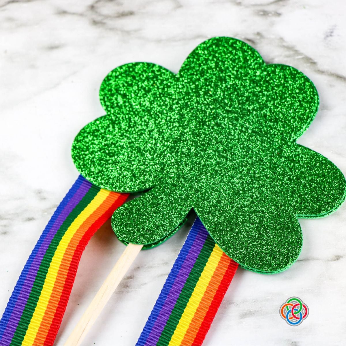 Attaching a second shamrock to a stick with ribbon strips for a shamrock wand craft | Irish American Mom Close-up of a glittery green foam shamrock being attached on top of ribbon streamers and a wooden dowel to finish a Saint Patrick's Day shamrock wand craft.