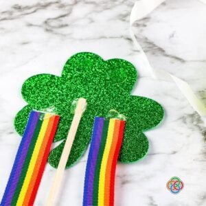 Attaching a stick and ribbon strips to a shamrock wand craft | Irish American Mom Close-up of a glittery green foam shamrock with rainbow ribbon strips and a wooden dowel attached using glue dots during a Saint Patrick's Day shamrock wand craft.