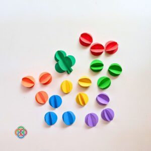 Multiple 3D paper shamrocks and colorful folded paper balls in red, orange, yellow, green, blue, and purple arranged on a white background for a Saint Patrick's Day garland craft, with Irish American Mom logo in the corner.