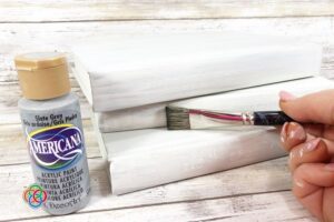 Creating a distressed look for a decorativ | Irish American Mom Close-up of a hand dry brushing grey acrylic paint onto white painted hardcover books to create a distressed farmhouse finish for a Saint Patrick's Day book stack craft.