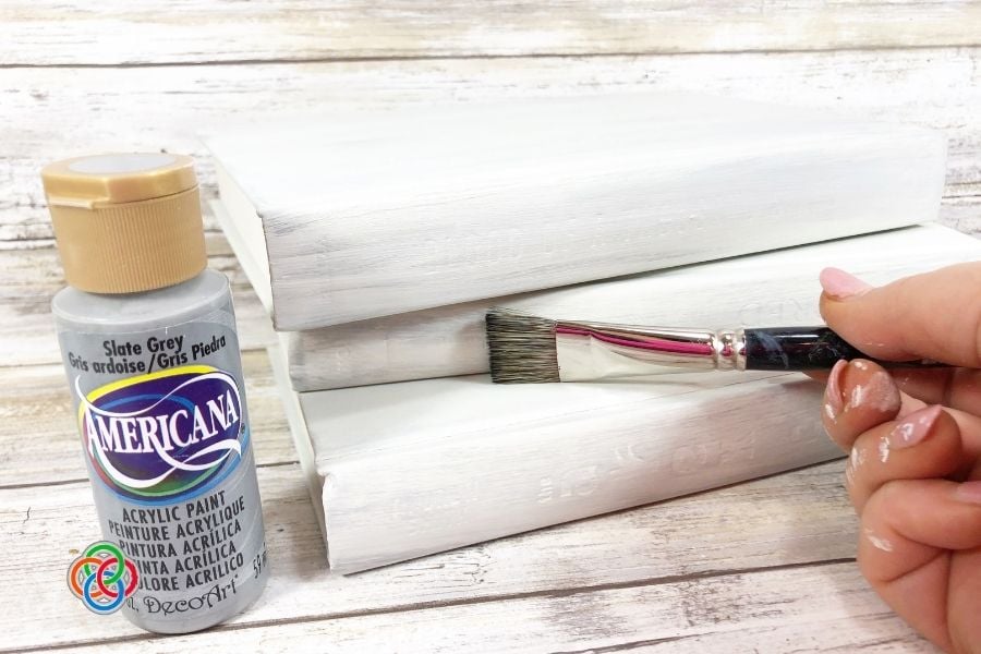 Creating a distressed look for a decorativ | Irish American Mom Close-up of a hand dry brushing grey acrylic paint onto white painted hardcover books to create a distressed farmhouse finish for a Saint Patrick's Day book stack craft.