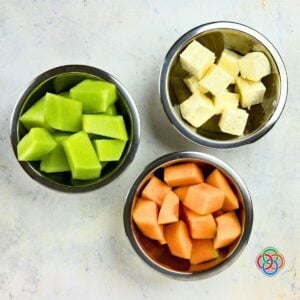 Overhead view of bowls filled with cubed honeydew melon, cubed cantaloupe, and cubed feta cheese prepared for Irish flag appetizer stacks.