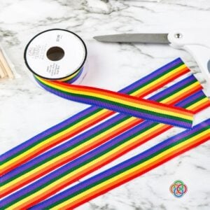 Cutting ribbon strips for a shamrock wand craft | Irish American Mom Close-up of a glittery green foam shamrock with rainbow ribbon strips and a wooden dowel attached using glue dots, shown during the assembly of a Saint Patrick's Day wand craft.