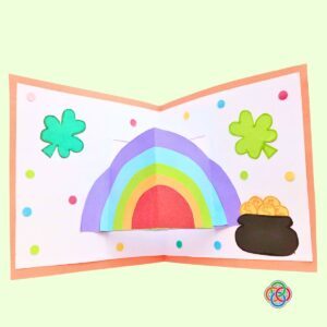 Decorate card with dots for Pop Up Rainbow Card Craft | Irish American Mom Completed Saint Patrick's Day pop up card decorated with colorful paper dots, featuring a 3D rainbow, shamrocks, and a pot of gold inside an orange-bordered card.