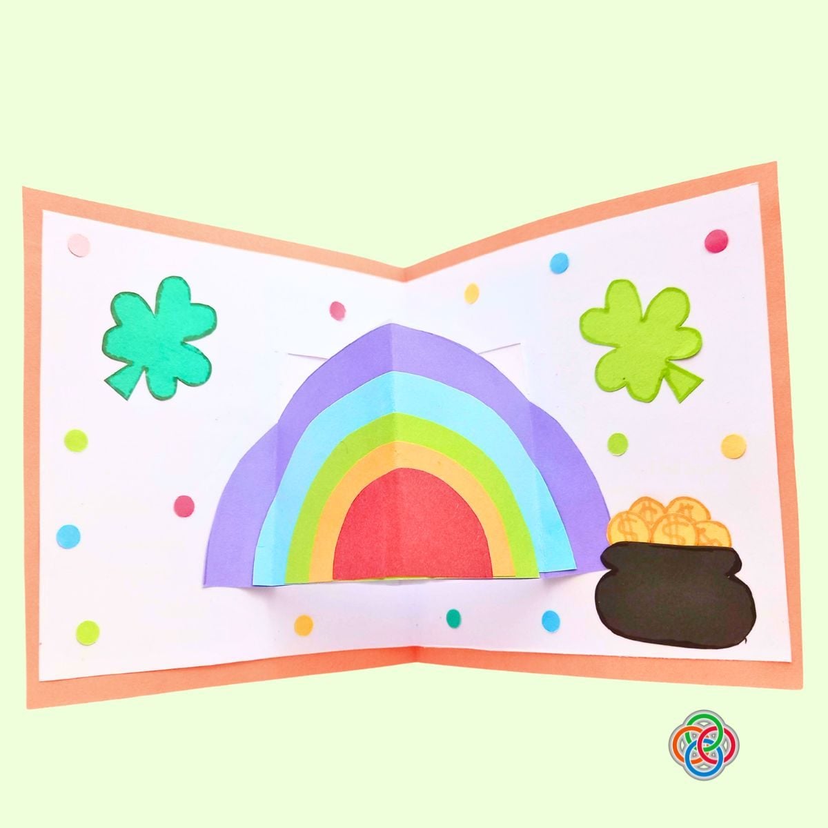 Decorate card with dots for Pop Up Rainbow Card Craft | Irish American Mom Completed Saint Patrick’s Day pop up card decorated with colorful paper dots, featuring a 3D rainbow, shamrocks, and a pot of gold inside an orange-bordered card.