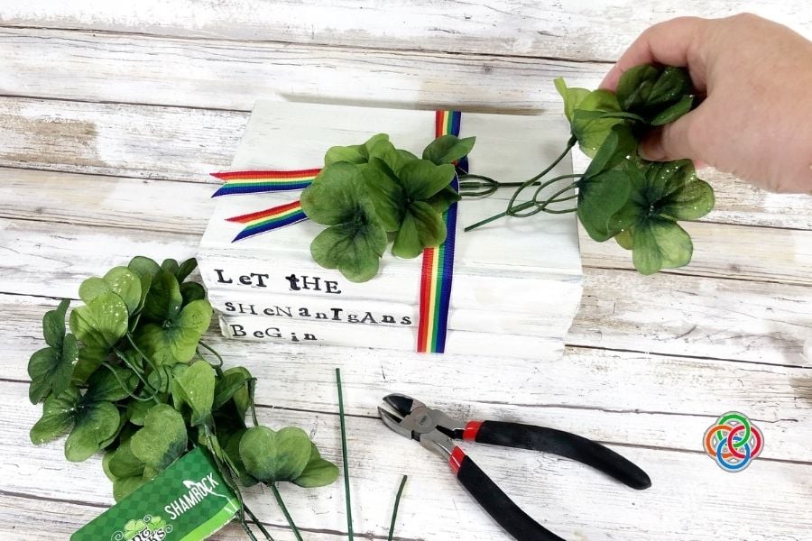 Decorating a book stack with shamrocks for | Irish American Mom Hand tucking green silk shamrocks under rainbow ribbon on a white painted “Let the Shenanigans Begin” book stack for a Saint Patrick’s Day farmhouse craft project.