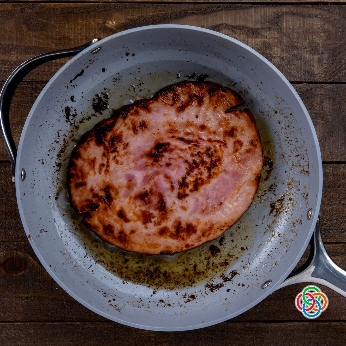 Overhead view of a ham steak frying in a skillet with browned edges and melted butter on a wooden table background.