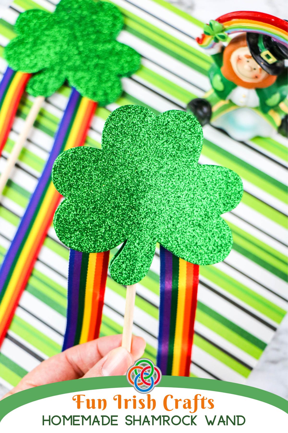 Fun Irish Crafts Homemade Shamrock Wand | Irish American Mom Hand holding a glittery green shamrock wand with rainbow ribbon streamers, displayed against a green striped background with Saint Patrick's Day decorations and Irish American Mom branding.