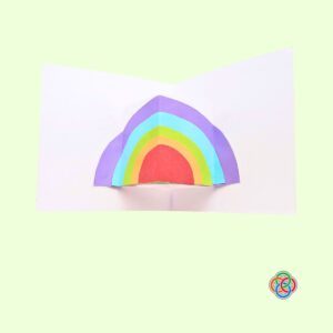 Glue the folded rainbow on the pop socket for Pop Up Rainbow Card Craft | Irish American Mom Layered paper rainbow glued onto the pop-up platform inside an open white Saint Patrick's Day card.