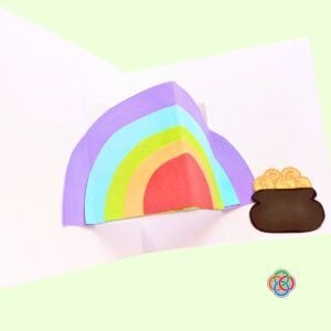 Glue the gold coins pot near the rainbow for Pop Up Rainbow Card Craft | Irish American Mom Open white pop up card featuring a layered 3D rainbow with a brown pot of gold glued beside it for a Saint Patrick's Day craft.