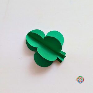Completed 3D green paper shamrock made by gluing three folded pieces together for a Saint Patrick's Day garland craft, with Irish American Mom logo in the corner.