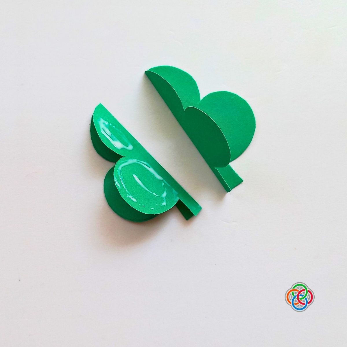 Two folded green shamrock paper pieces being glued together to create a 3D shamrock for a Saint Patrick's Day garland craft, with Irish American Mom logo in the corner.