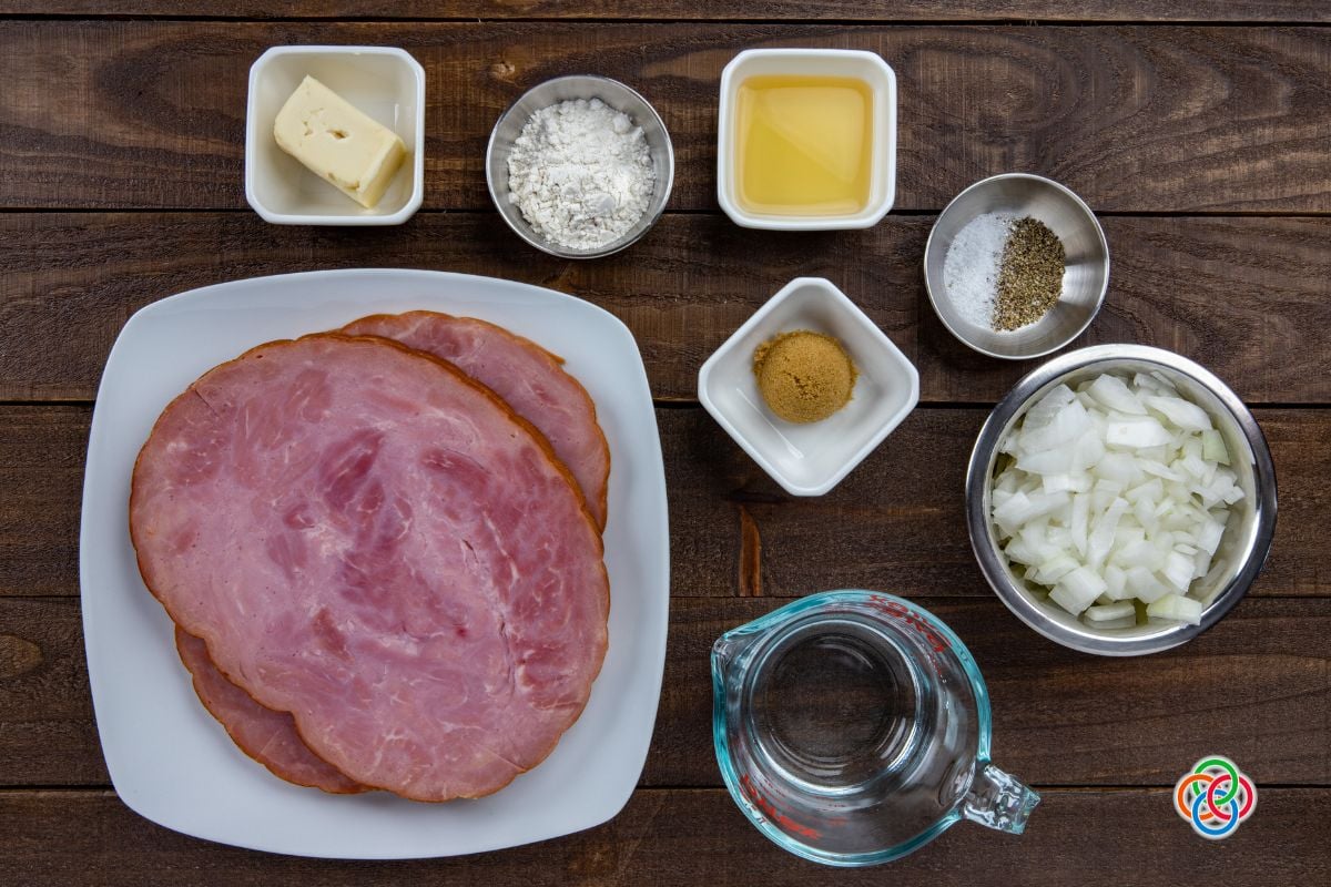 Overhead view of ingredients for ham steaks with whiskey sauce including ham slices, chopped onion, butter, flour, brown sugar, whiskey, salt, pepper, and water on a wooden table.