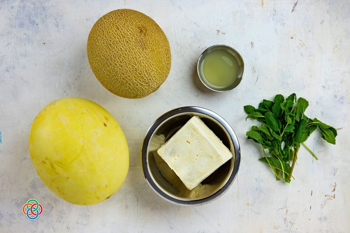 Overhead view of ingredients for Irish flag appetizer stacks including whole honeydew and cantaloupe melons, a block of feta cheese in a bowl, fresh mint sprigs, and a small bowl of lime juice on a light surface.