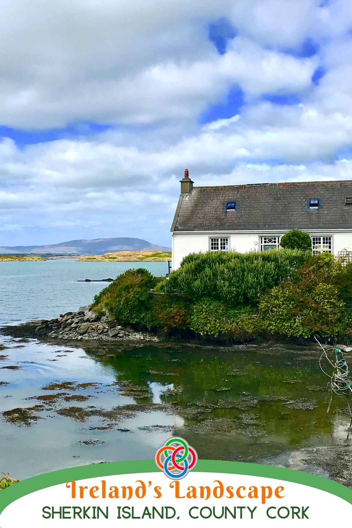 White cottage overlooking calm coastal waters on Sherkin Island, County Cork, Ireland, with rocky shoreline, green shrubs, and distant hills beneath a cloudy blue sky.