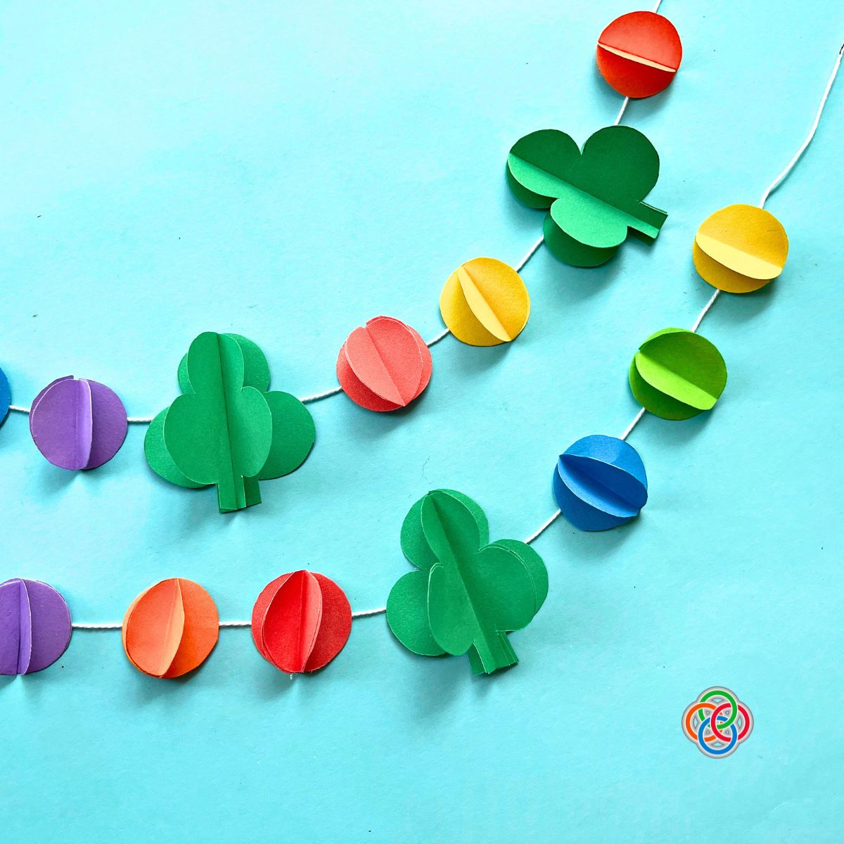 Colorful Saint Patrick’s Day paper garland with 3D green shamrocks and rainbow paper ball shapes strung on white string against a blue background, with Irish American Mom