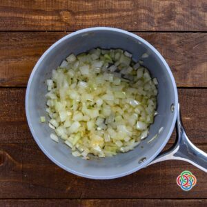 Overhead view of chopped onions sautéing in a saucepan with butter on a wooden surface, softening and turning translucent.