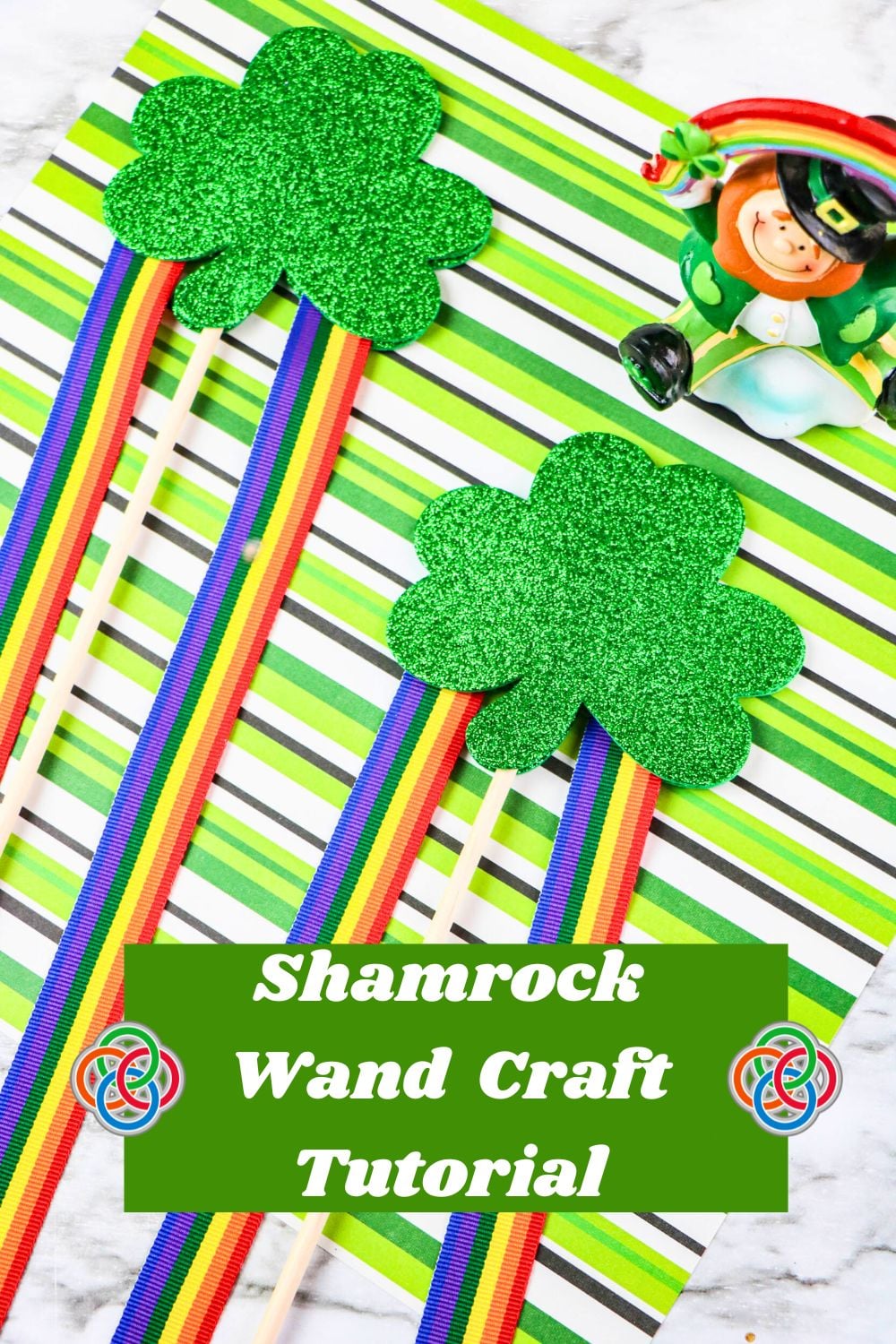 Shamrock Wand Craft Tutorial | Irish American Mom Two glittery green shamrock wands with rainbow ribbon streamers on a green striped background, featuring the text