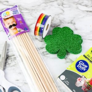 Supplies for a shamrock wand craft | Irish American Mom Flat lay of shamrock wand craft supplies including glittery foam shamrock cutouts, rainbow ribbon roll, wooden dowels, glue dots, and scissors on a marble surface.