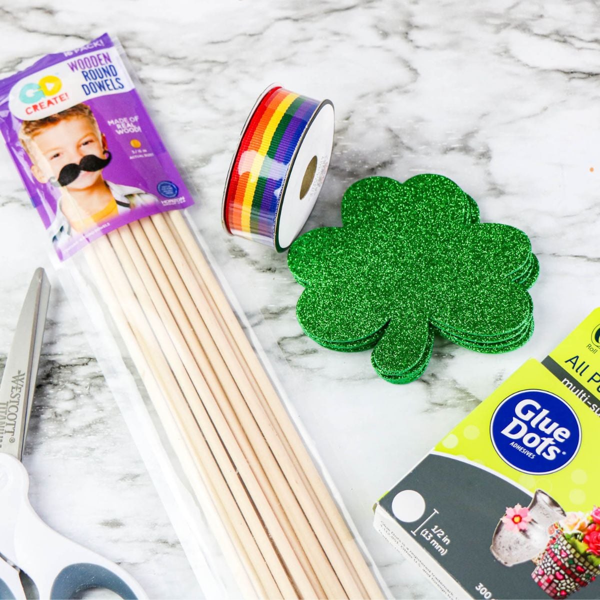 Supplies for a shamrock wand craft | Irish American Mom Flat lay of shamrock wand craft supplies including glittery foam shamrock cutouts, rainbow ribbon roll, wooden dowels, glue dots, and scissors on a marble surface.
