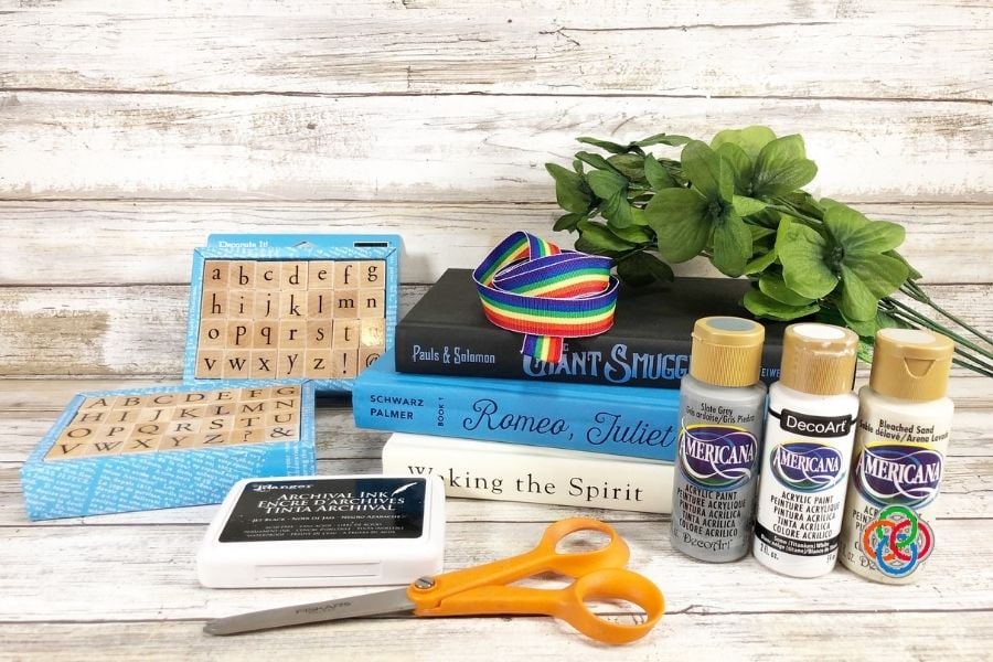 Supplies required for a book stack craft wi | Irish American Mom Craft supplies laid out for a Saint Patrick’s Day book stack project including hardcover books, rainbow ribbon, silk shamrocks, alphabet stamps, ink pad, craft paint bottles, and scissors.