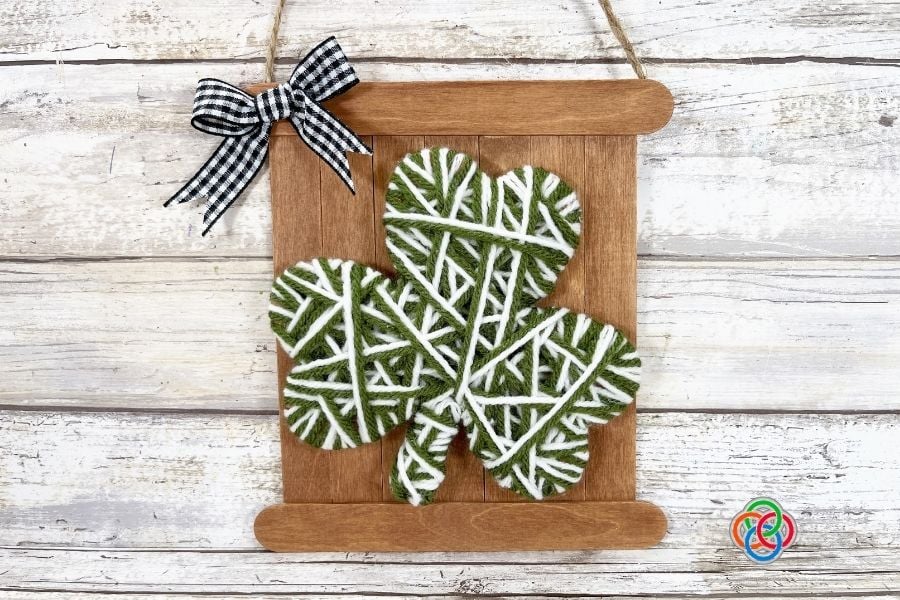 Finished yarn wrapped shamrock mounted on a rustic craft stick sign with a black gingham bow and twine hanger for a St. Patrick’s Day decoration.