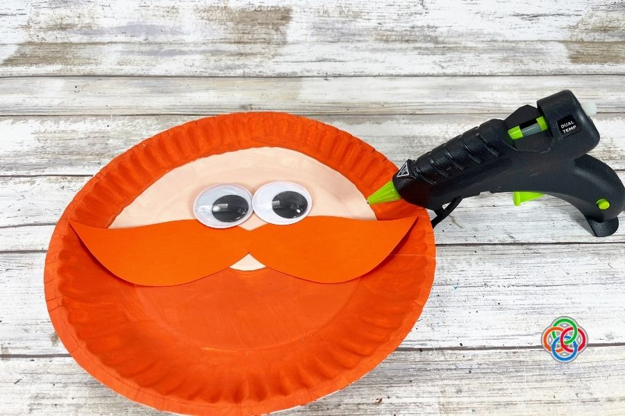 Paper plate leprechaun craft with orange beard, googly eyes, and cardstock mustache being assembled using a hot glue gun during a St. Patrick’s Day kids craft tutorial.