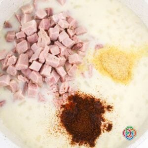 Diced ham, garlic powder, and smoked paprika being added to a creamy potato soup base in a pot.