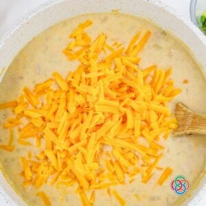 Shredded cheddar cheese being added to a creamy ham and potato soup in a pot, ready to melt into a rich and cheesy finish.