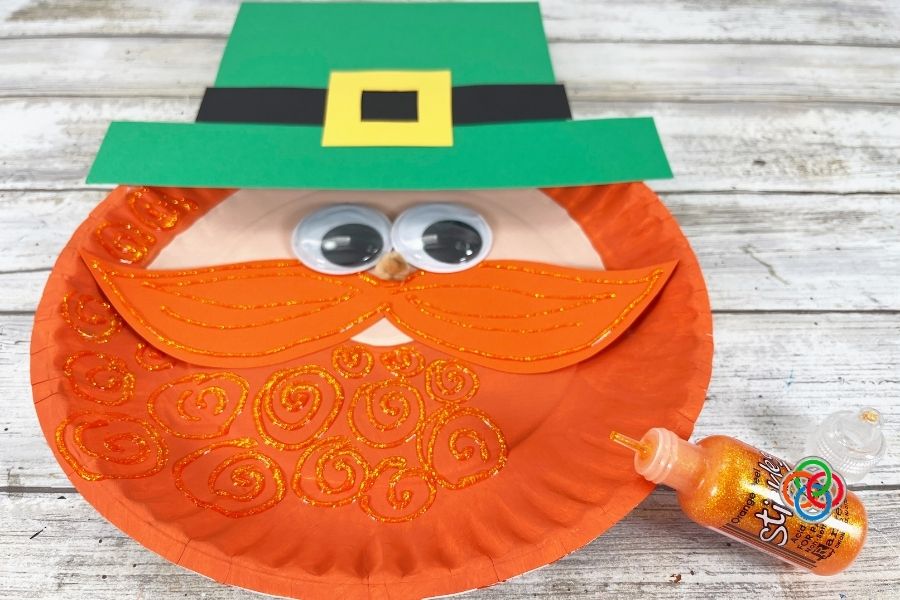 Paper plate leprechaun craft with orange beard and mustache decorated with glitter glue swirls while adding sparkle details during a St. Patrick’s Day kids craft tutorial.