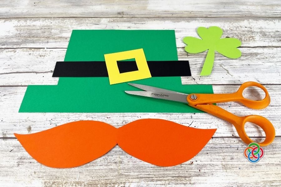 Cut cardstock pieces for a paper plate leprechaun craft including green hat, black hat band, yellow buckle, shamrock, and orange mustache with scissors on a wooden background.