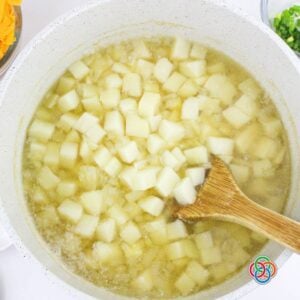 Diced potatoes simmering in a pot of broth with a wooden spoon, forming the base of a creamy ham and potato soup.