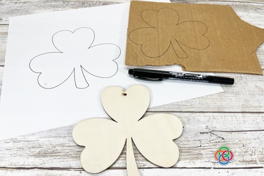 Printable shamrock template traced onto cardboard with marker and scissors preparing a shamrock shape for a St. Patrick’s Day yarn craft project.