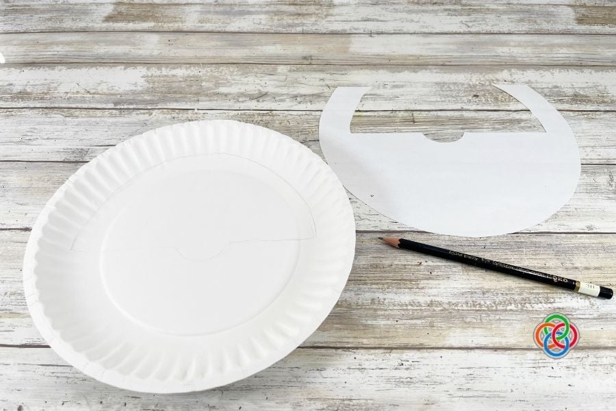Paper plate with leprechaun beard template traced onto it next to the printed pattern and pencil during a paper plate leprechaun craft tutorial for kids.