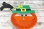 Finished paper plate leprechaun craft with glittery orange beard, googly eyes, green hat with yellow buckle, and shamrock decoration during a St. Patrick’s Day kids craft tutorial.