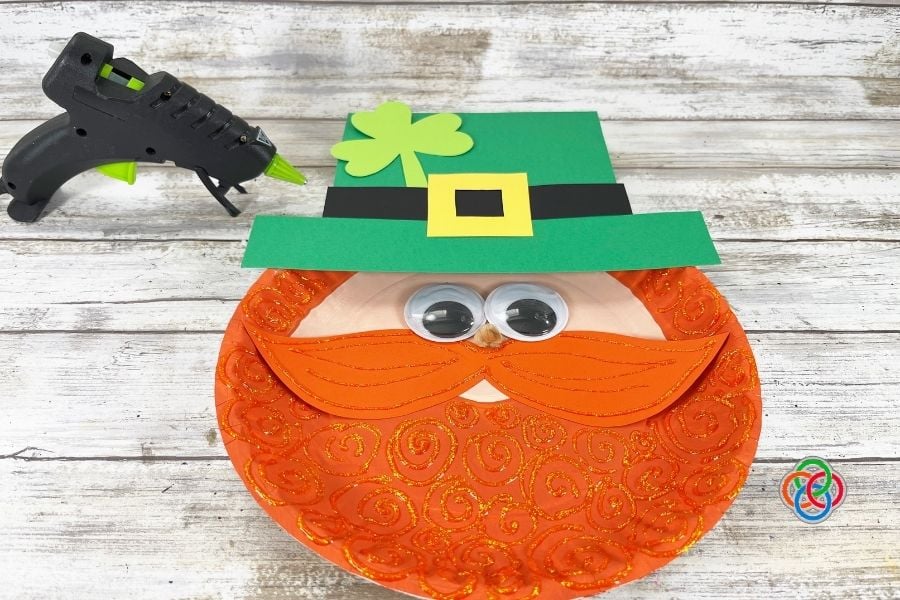 Finished paper plate leprechaun craft with glittery orange beard, googly eyes, green hat with yellow buckle, and shamrock decoration during a St. Patrick’s Day kids craft tutorial.