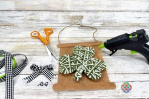 Yarn wrapped shamrock glued onto a rustic craft stick sign with ribbon bow, scissors, and glue gun during a St. Patrick's Day craft project.