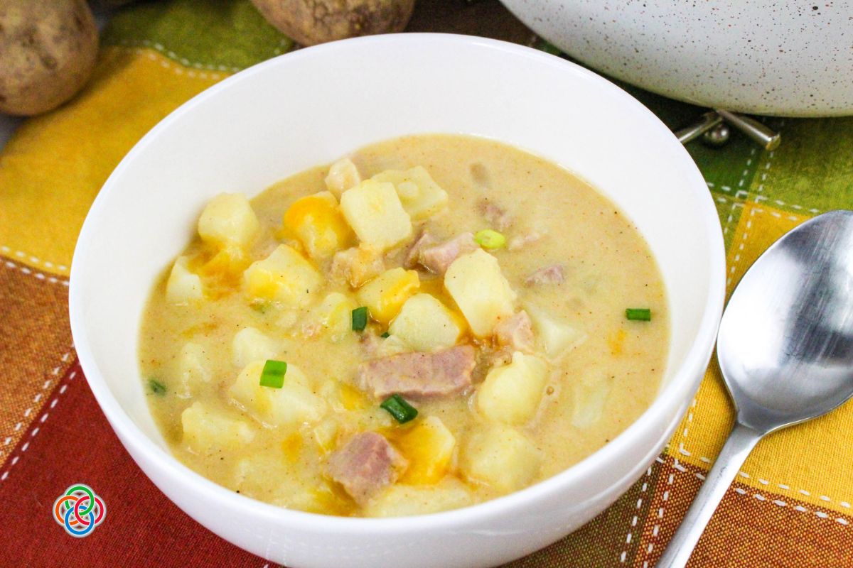 Bowl of creamy cheddar ham and potato soup with chunks of potato, ham, melted cheese, and green onions, served on a table with a spoon beside it.