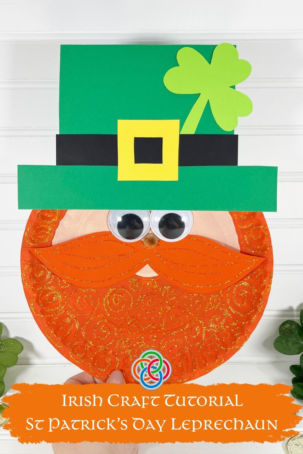 Finished paper plate leprechaun craft with glittery orange beard, googly eyes, green hat with yellow buckle, and shamrock decoration during a St. Patrick’s Day kids craft tutrial. with text banner.