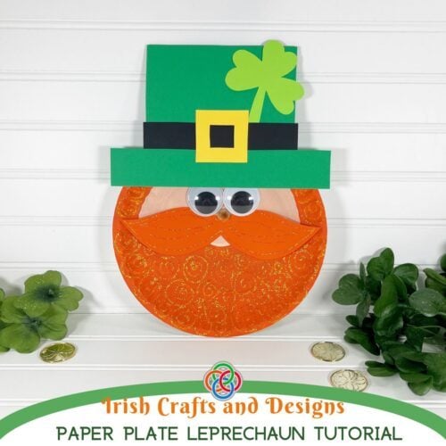 Leprechaun paper plate craft with glittery orange beard, googly eyes, green hat with yellow buckle, and shamrock decoration for a St. Patrick's Day kids craft tutorial.