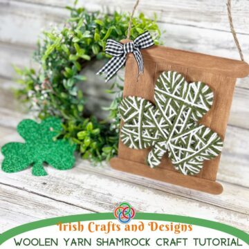 Yarn wrapped green and white shamrock mounted on a rustic craft stick sign with a gingham bow and twine hanger beside a wreath for a St. Patrick’s Day craft display.